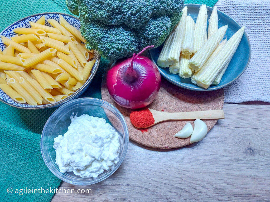 Ingredients lined up on a background of green and beige table cloth, from the left, a bowl of uncooked pasta, bowl of cottage cheese, head of broccoli, red onion, a wooden teaspoon with paprika powder, baby corn on a blue plate and two cloves of garlic.