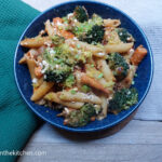 On a wooden table, with a beige table cloth and a green napkin, a blue plate with vegetable and cottage cheese pasta.