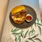 On a silver coloured table, a cloth napkin with a green leaf pattern, and a black plate with a barbecue hamburger on a brioche bun, with bacon, cheese and lettuce visible, next to a small white bowl with barbecue sauce.