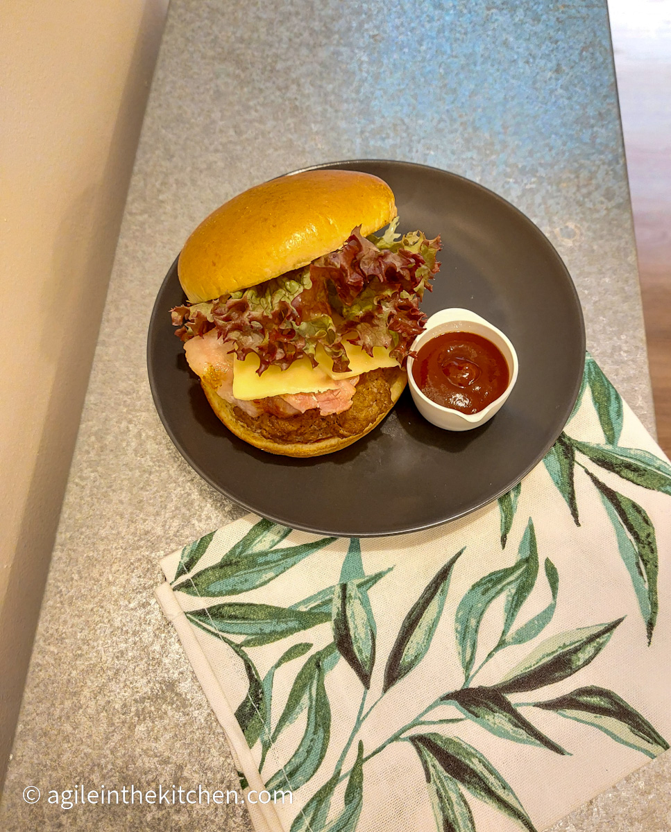 On a silver coloured table, a cloth napkin with a green leaf pattern, and a black plate with a barbecue hamburger on a brioche bun, with bacon, cheese and lettuce visible, next to a small white bowl with barbecue sauce.