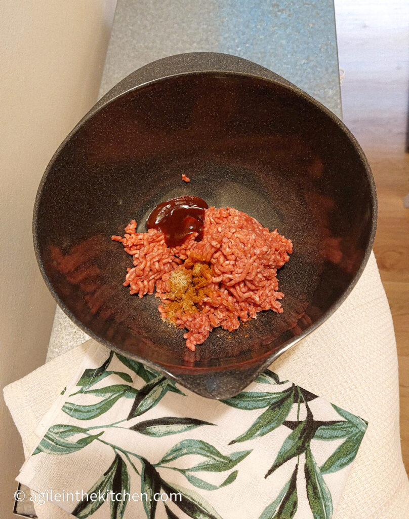 On a silver coloured table, a cloth napkin with a green leaf pattern, and a black plastic mixing bowl filled with mince meat, barbecue sauce and spice mix.