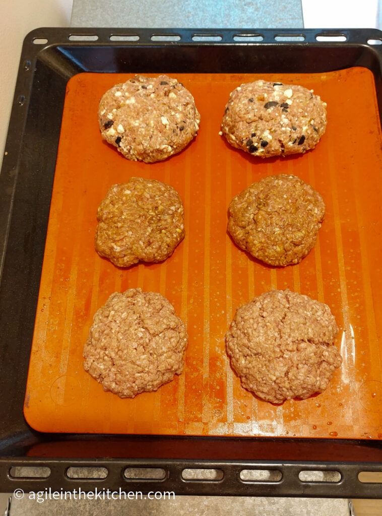 On a silver coloured table, a black cookie sheet with a red silicone cookie sheet cover, six raw hamburgers in two columns with different flavours: Mediterranean with feta cheese and black olives, barbecue with barbecue sauce and spices, Asian with soya sauce, sesame oil and ginger