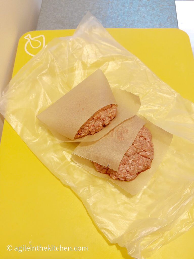 On a silver coloured table, a yellow, plastic cutting board, two raw hamburgers wrapped in baking paper on top of a plastic bag, ready to be frozen.