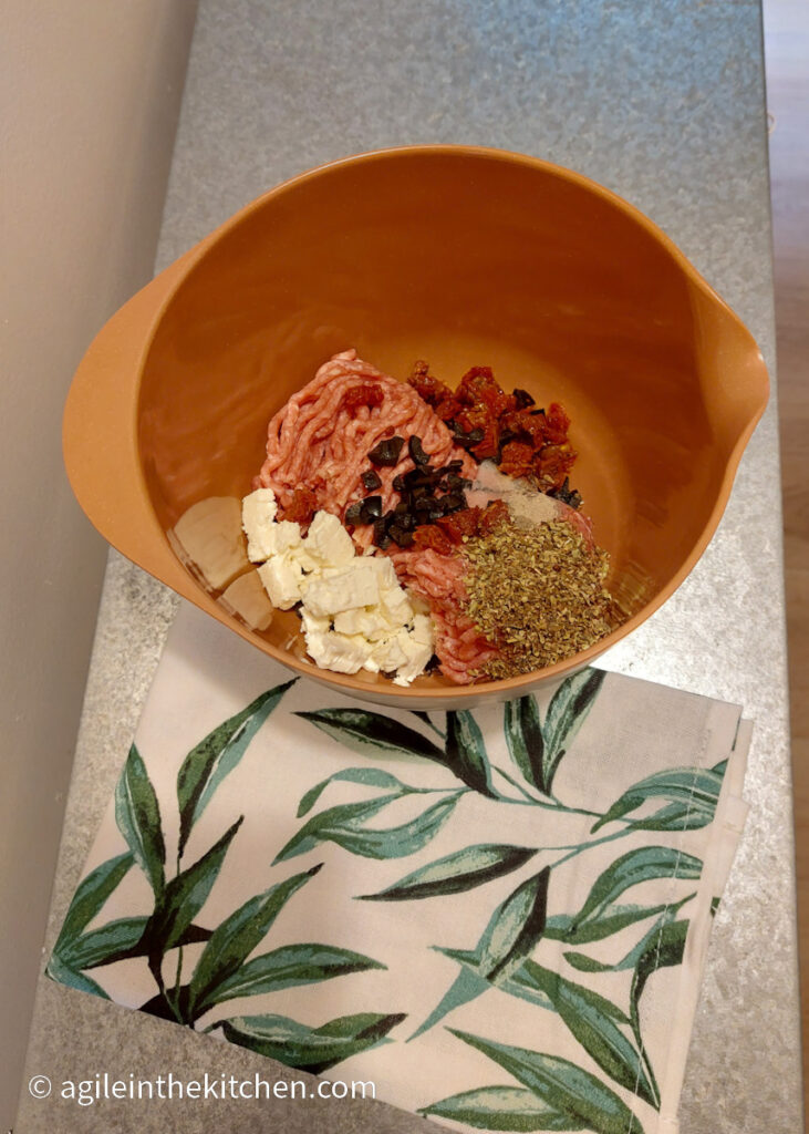 On a silver coloured table, a white cloth napkin with a green leaf pattern, an orange, plastic bowl with mince meat, chopped feta cheese, sun-dried tomatoes and black olives, as well as heaps of dried oregano.