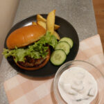 On a silver coloured table with a folded pink gingham cloth a glass bowl with tzatziki and a black plate with a Mediterranean hamburger dressed with leafy lettuce, three cucumber slices on the side and a few potato wedges.