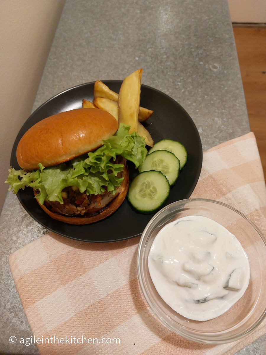 On a silver coloured table with a folded pink gingham cloth a glass bowl with tzatziki and a black plate with a Mediterranean hamburger dressed with leafy lettuce, three cucumber slices on the side and a few potato wedges.