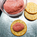 On a silver coloured table, a glass bowl with pink beet hummus, two whole wheat crackers on the side. In the foreground a wheat cracker with a dollop of pink beet hummus