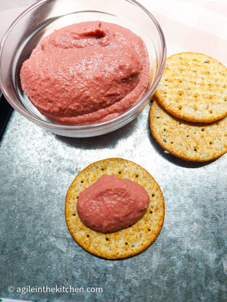 On a silver coloured table, a glass bowl with pink beet hummus, two whole wheat crackers on the side. In the foreground a wheat cracker with a dollop of pink beet hummus