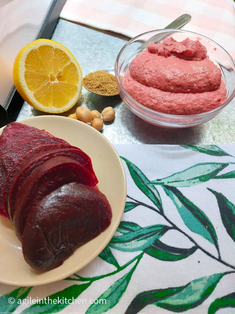 On a silver coloured table with a white table cloth with a green leaf pattern: a small plate with sliced, cooked beet root, a half lemon, some chickpeas, a table spoon with cumin and a glass bowl with pink beet hummus.