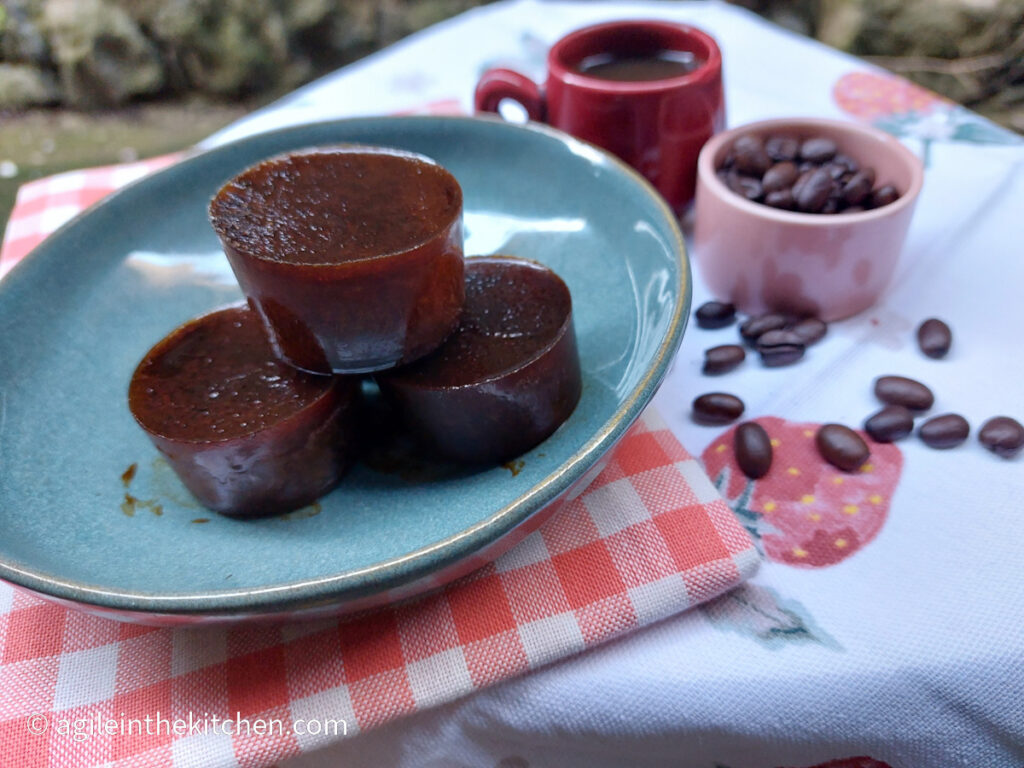 On a white table cloth with a strawberry pattern, a red folded gingham cloth, a blue plate with three large coffee ice cubes. In the background a red espresso cup and a pink small bowl with coffee beans, a few coffee beans have spilled out from the small bowl.