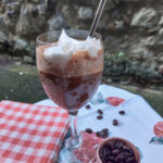 On a white table cloth with a strawberry pattern, a red folded gingham cloth, a glass with a stem and a printed pattern, filled with blended ice coffee and topped with whipped cream and a silver coloured reusable straw, a pink small bowl with coffee beans, a few coffee beans have spilled out from the small bowl.