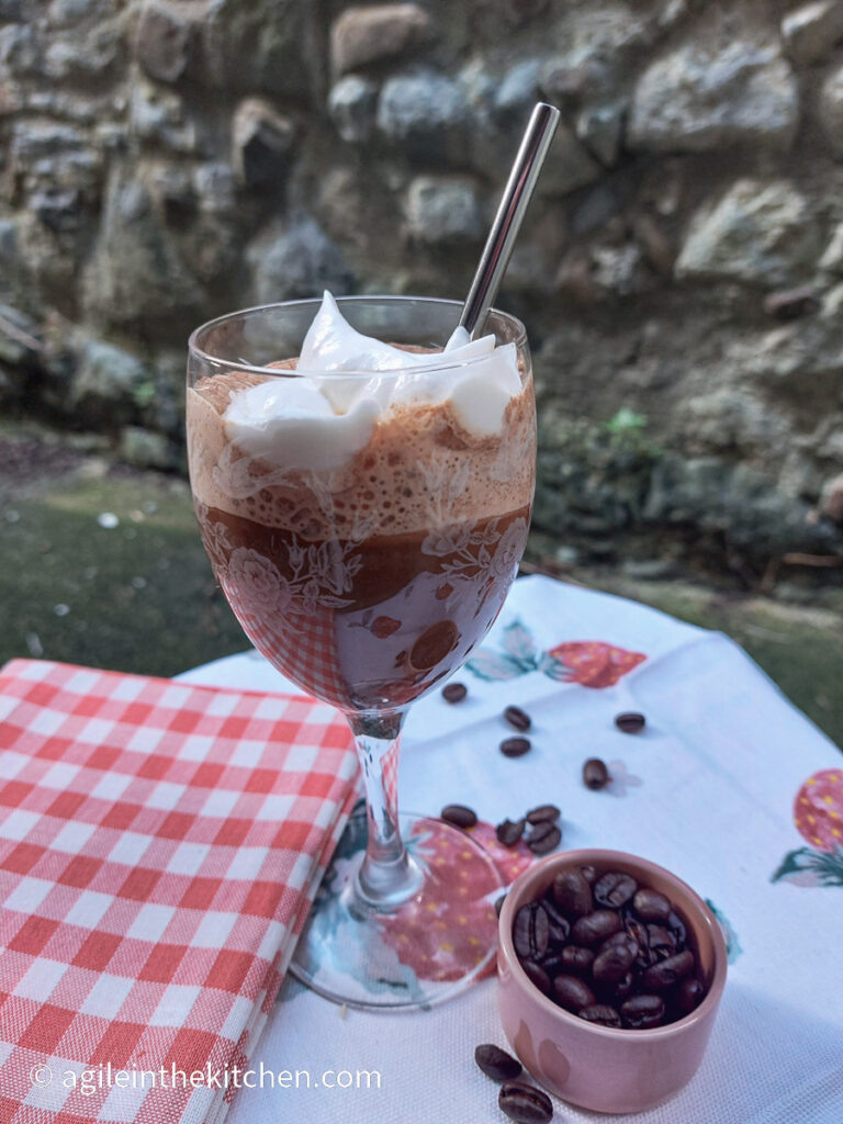 On a white table cloth with a strawberry pattern, a red folded gingham cloth, a glass with a stem and a printed pattern, filled with blended ice coffee and topped with whipped cream and a silver coloured reusable straw, a pink small bowl with coffee beans, a few coffee beans have spilled out from the small bowl.