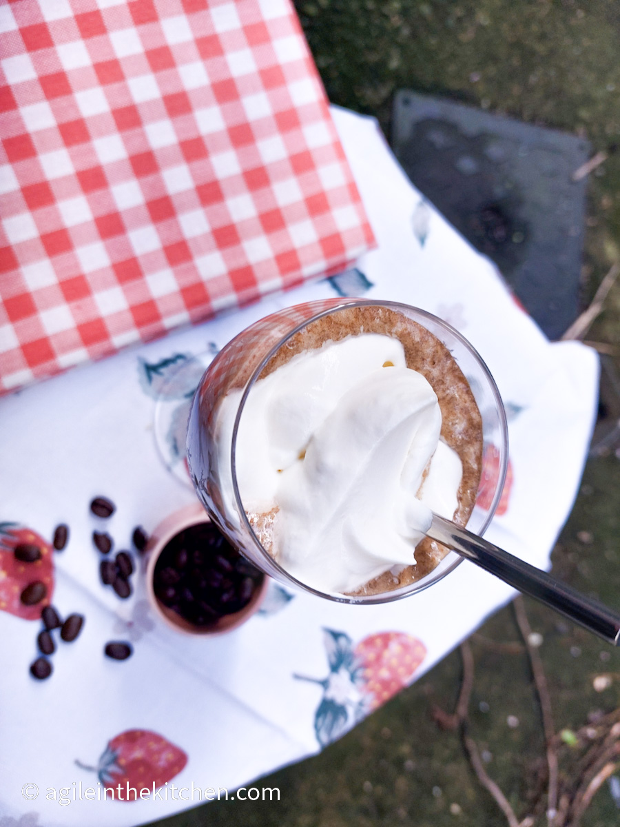 A photo taken from above. On a white table cloth with a strawberry pattern, a red folded gingham cloth, a glass with a stem and a printed pattern, filled with ice coffee topped with whipped cream, and a silver coloured reusable straw, a pink small bowl with coffee beans, a few coffee beans have spilled out from the small bowl.