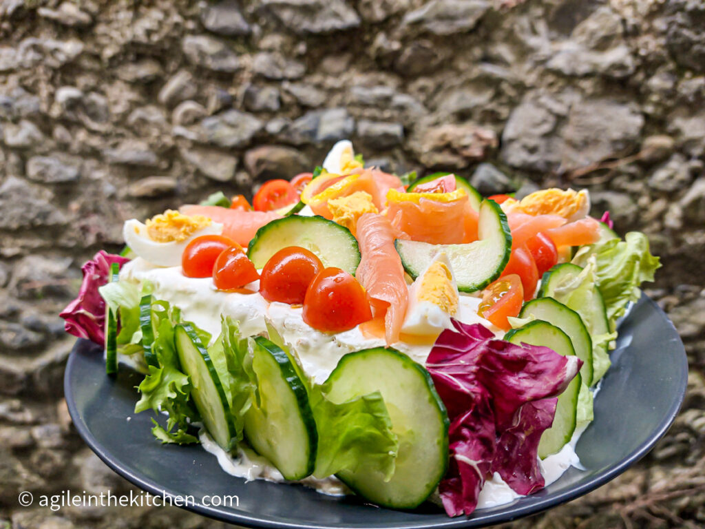 On a stone wall background, a whole smörgåstårta on a black matte plate, on the edges sliced fresh cucumber and lettuce leaves, on top sliced cocktail tomatoes, sliced fresh cucumber, quartered boiled eggs and slices of smoked salmon