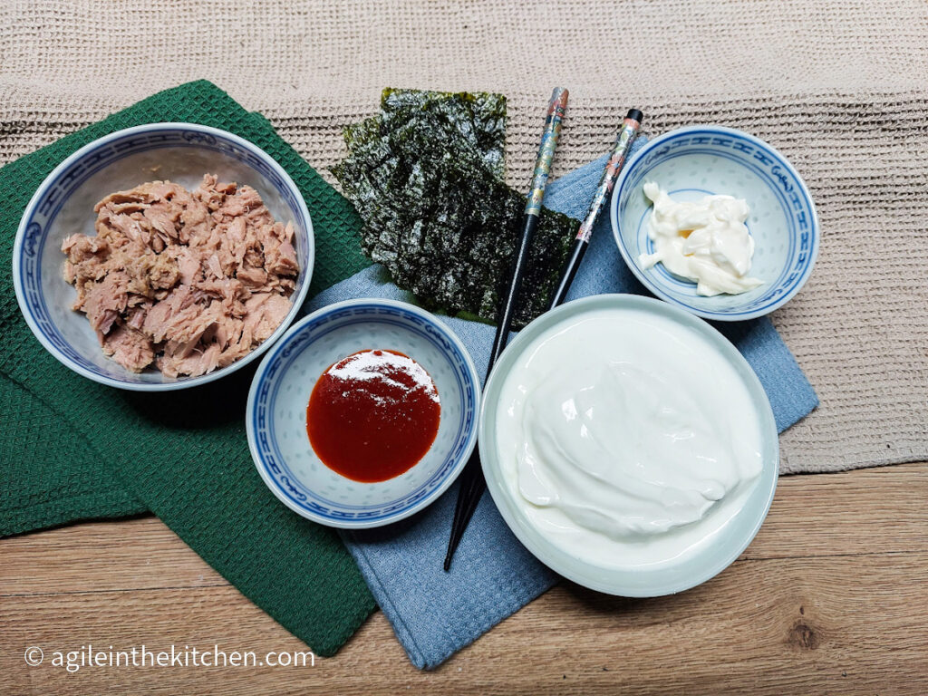 On a beige table cloth, two folded cloth napkins, one dark green and one light blue, four bowls are lined up with ingredients to make spicy tuna bowl, tuna, mayonnaise, plain yogurt and shiraza, a pair of black chopsticks and some nori sheets are in the middle