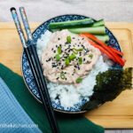 On a beige table cloth, a wooden cutting board two folded cloth napkins, one dark green and one light blue. A porcelain bowl with a blue pattern filled with sushi rice, spicy tuna, sliced cucumber and red peppers, a sheet of nori and two chopsticks balancing on top.