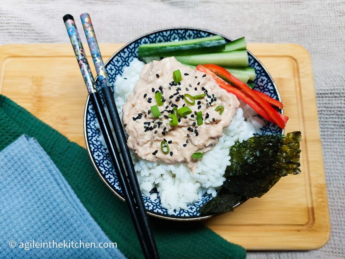 On a beige table cloth, a wooden cutting board two folded cloth napkins, one dark green and one light blue. A porcelain bowl with a blue pattern filled with sushi rice, spicy tuna, sliced cucumber and red peppers, a sheet of nori and two chopsticks balancing on top.