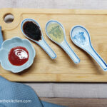 On a beige table cloth, a wooden chopping board, a folded blue cloth napkin in the lower left corner. On top of the wooden chopping board, a small bowl of Shiraza, three ceramic spoons with soy sauce, sesame oil and rice vinegar.