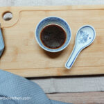 On a beige tablecloth, a wooden chopping board, a folded blue cloth napkin in the lower left corner. On top of the wooden chopping board, a small bowl with mixed Asian dipping sauce and a ceramic spoon laying next to it.