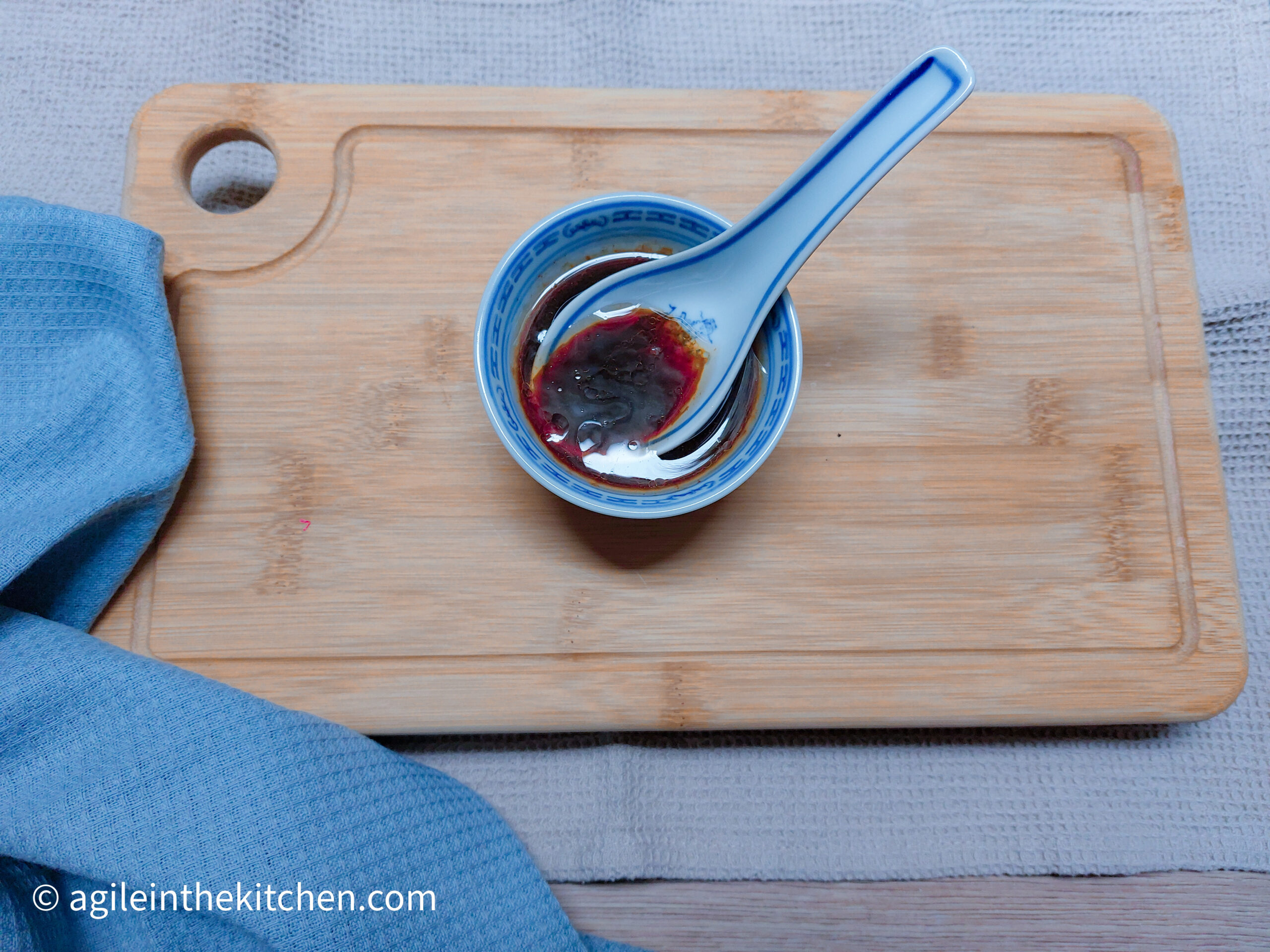 On a beige tablecloth, a wooden chopping board, a folded blue cloth napkin in the lower left corner. On top of the wooden chopping board, a small bowl with mixed Asian dipping sauce and a ceramic spoon.