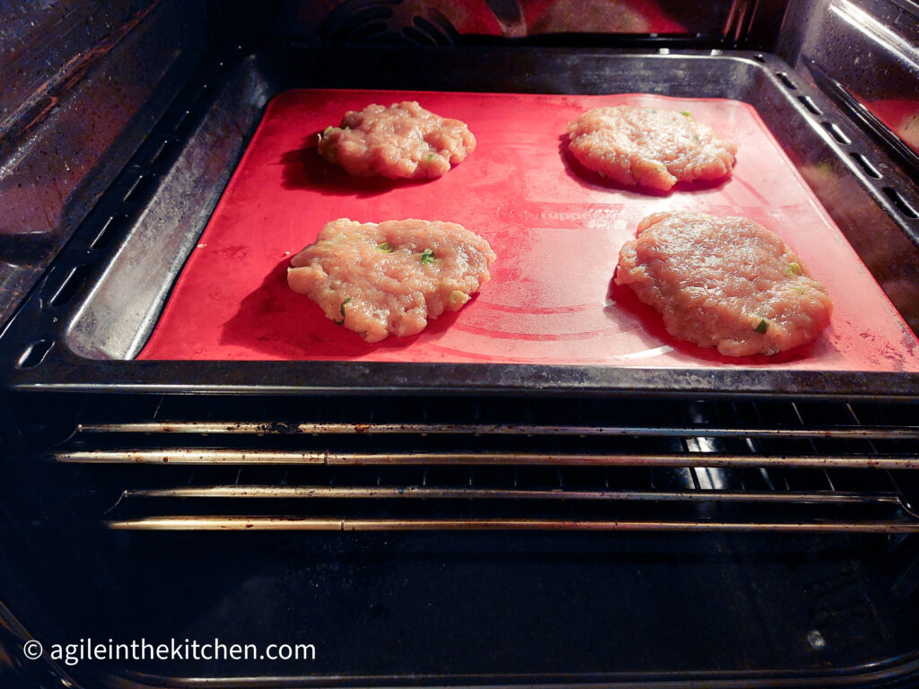 Inside of an oven: a cookie sheet, with a red silicone baking mat, four, raw, Asian turkey hamburgers.