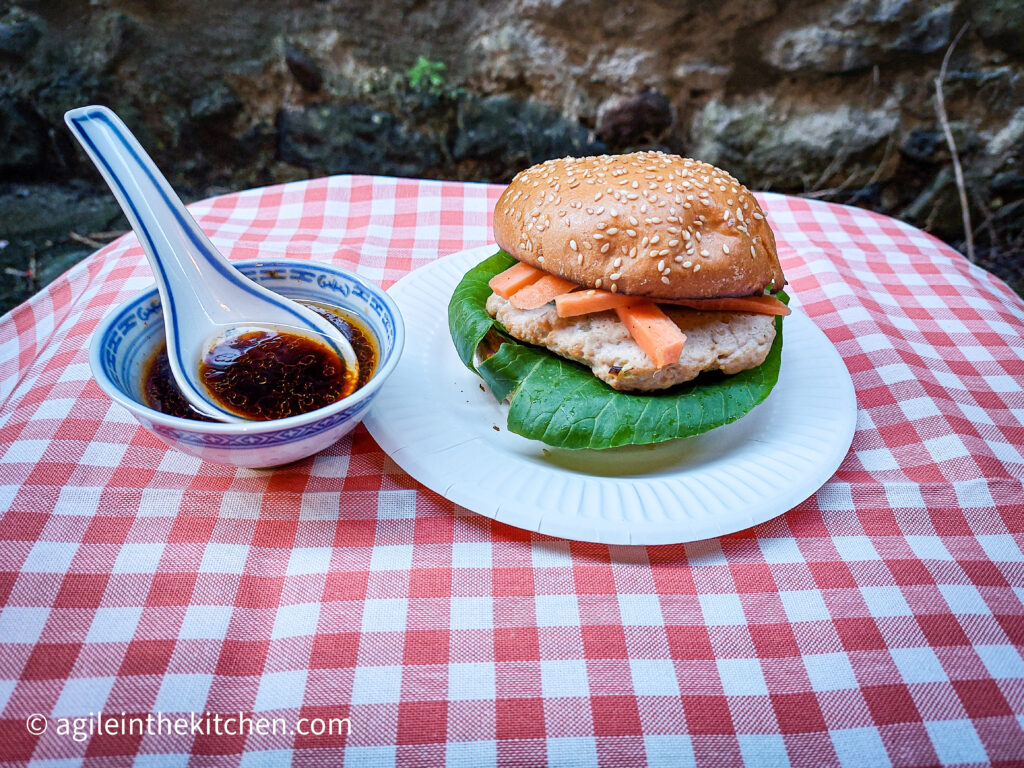 On a red gingham table cloth a paper plate with an Asian hamburger dressed with Pok Choi and pickled carrots. Next to it a small porcelain bowl, with a porcelain spoon, containing an Asian dipping sauce