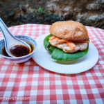 On a red gingham table cloth a paper plate with an Asian hamburger dressed with Pok Choi and pickled carrots. Next to it a small porcelain bowl, with a porcelain spoon, containing an Asian dipping sauce