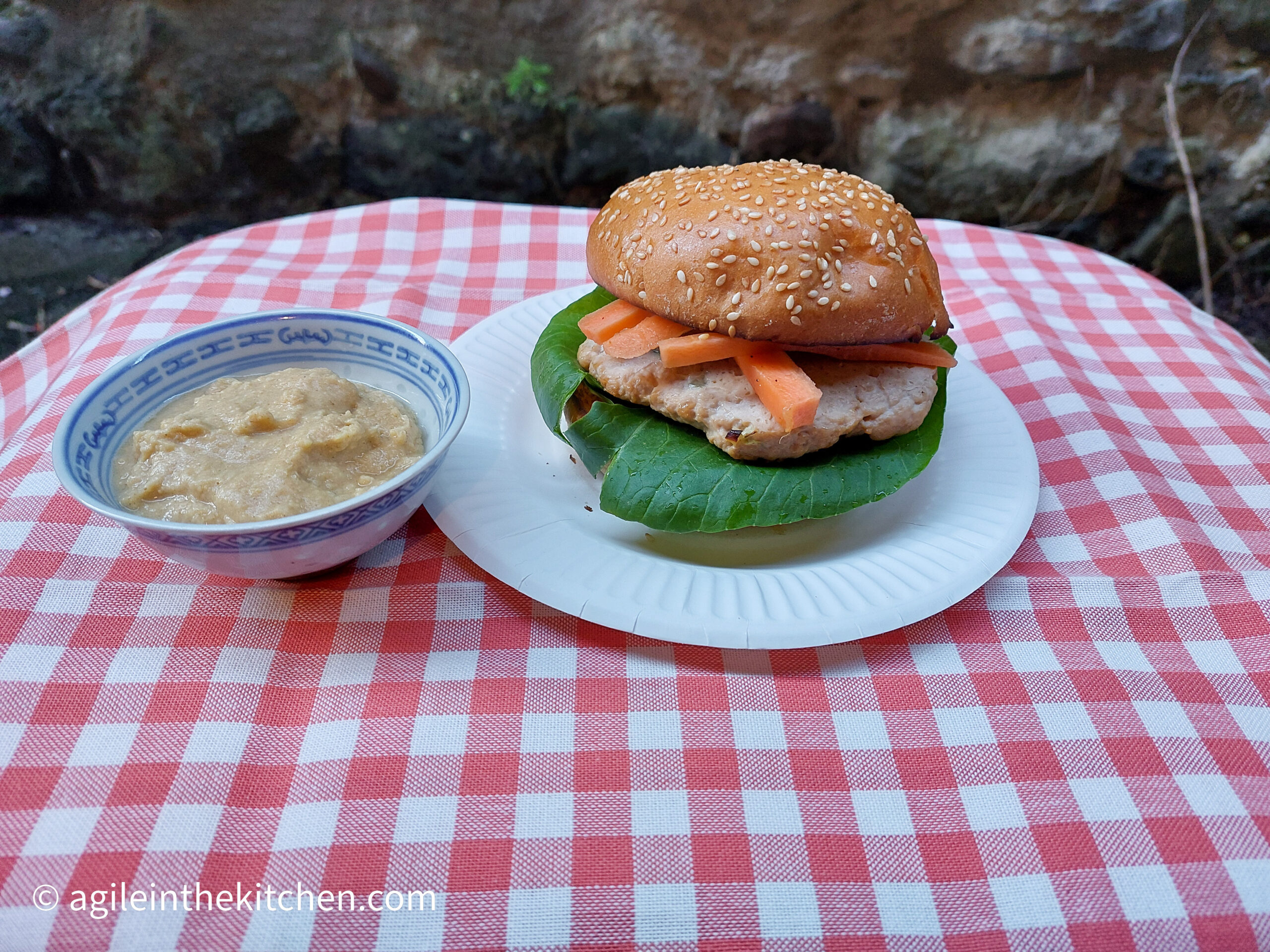 On a red gingham table cloth a paper plate with an Asian hamburger dressed with Pok Choi and pickled carrots. Next to it a small porcelain bowl, with a wasabi hummus.