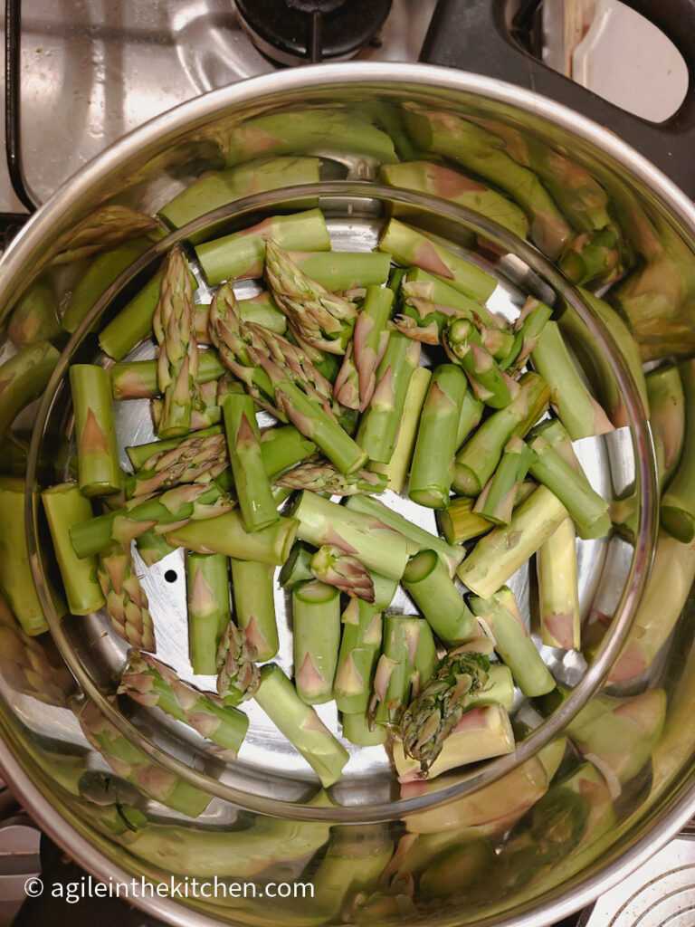 In a silver steamer, green asparagus are cut into smaller pieces.