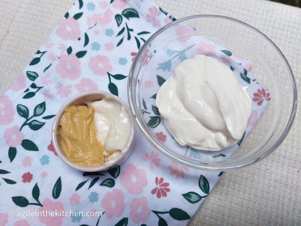 On a white table cloth, a folded cloth napkin with a flower and leaf pattern, a glass bowl with Turkish yogurt, a smaller pot with half Dijon mustard and half mayonnaise.