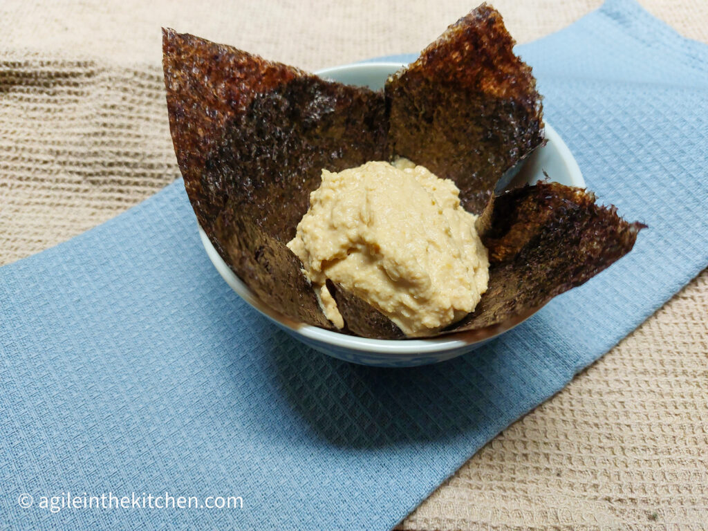 On a beige table cloth background, a folded light blue cloth napkin, a porcelain bowl with a nori sheet and a large portion of hummus wasabi