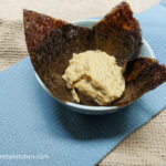 On a beige table cloth background, a folded light blue cloth napkin, a porcelain bowl with a nori sheet and a large portion of hummus wasabi