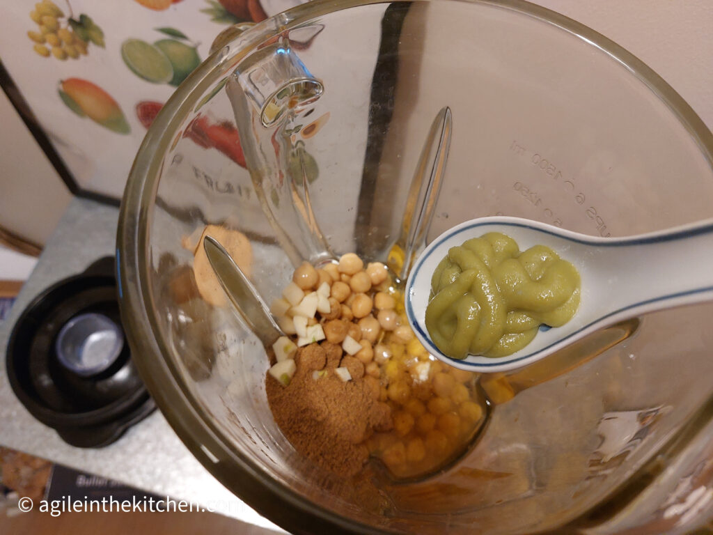 From above, a blender with hummus ingredients, chopped garlic, chickpeas, cumin, olive oil, a porcelain spoon with wasabi is about to be added.