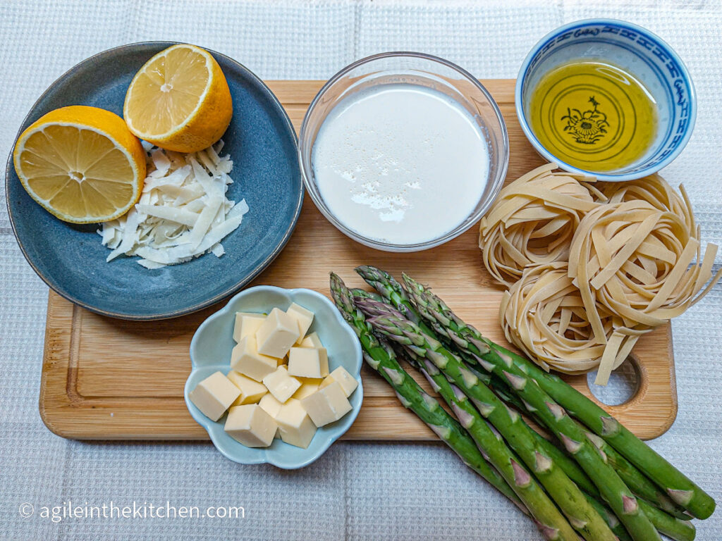 On a white table cloth, a cutting board with ingredients to make lemon asparagus pasta. Clockwise: a lemon cut in half on a blue plate together with shredded parmesan cheese, a glass bowl with heavy cream, a porcelain bowl with olive oil, a few nests of tagliatelle, asparagus, a blue flower shaped bowl with butter cut up in cubes