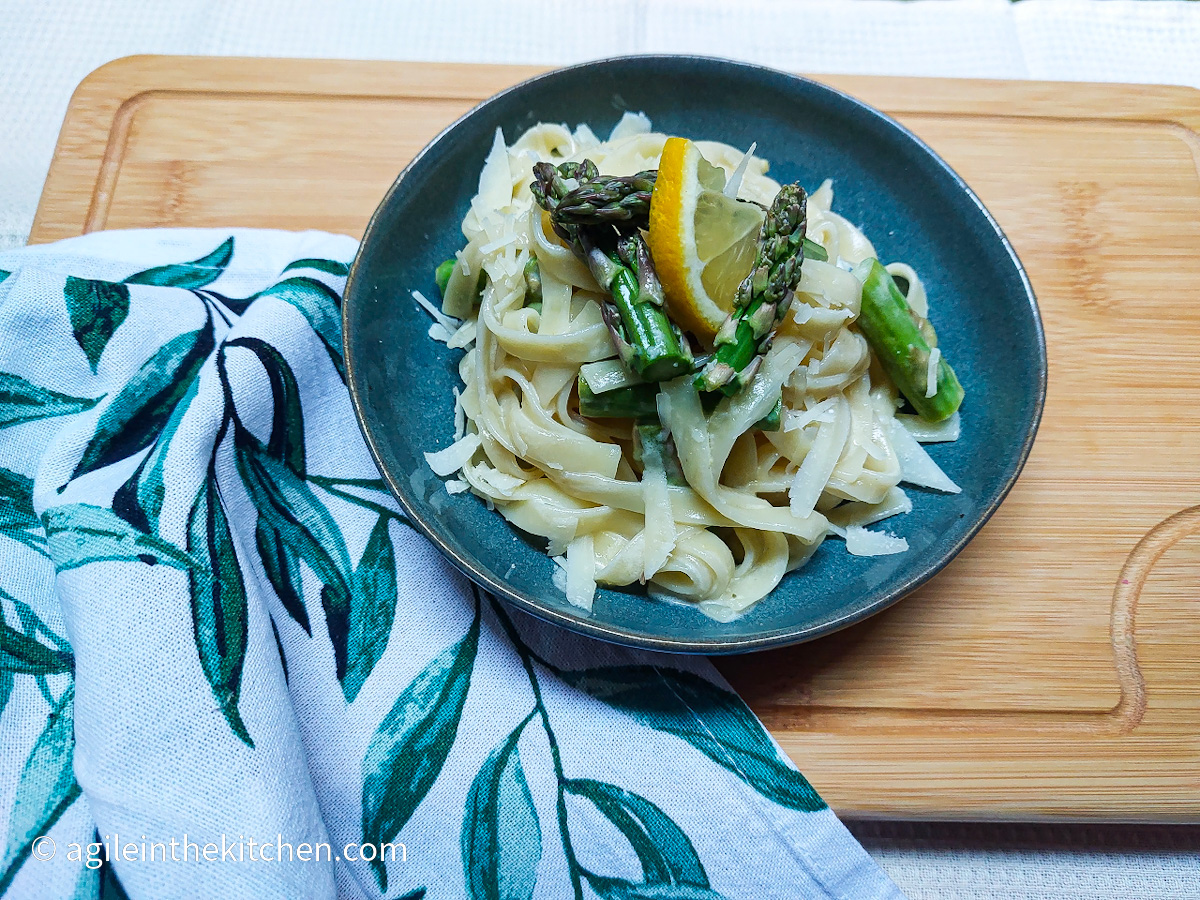 On a cutting board, a white cloth napkin with a green leaf pattern and a blue plate with lemon asparagus pasta laid out.