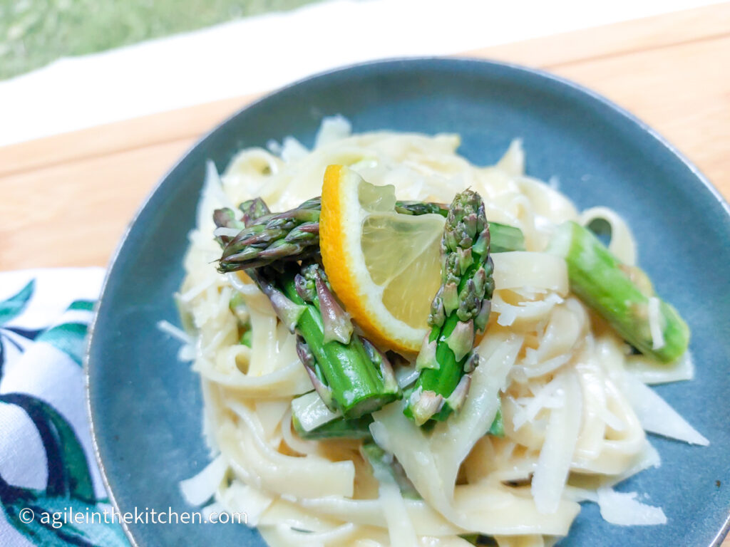 A blue plate with lemon asparagus pasta laid out. In focus is a few pieces of cut asparagus and a quarter slice of lemon.