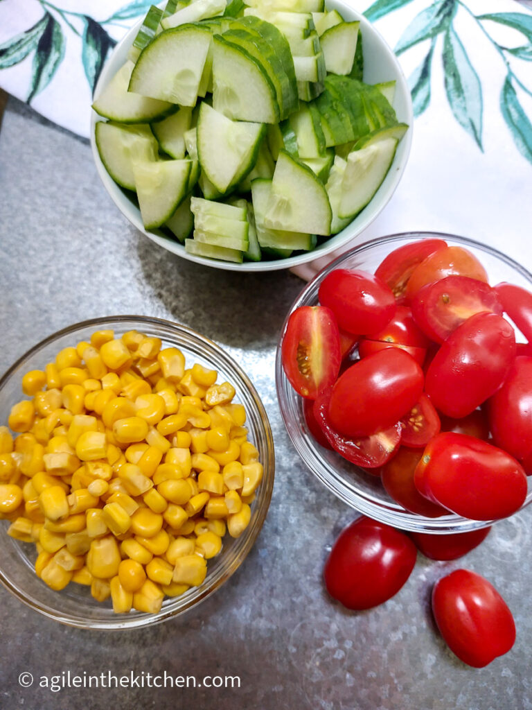 On a silver back ground with a white table cloth with a green leaf pattern in the upper half, three bowls of ingredients to make a basic pasta salad, cucumber cut in quarters, baby plum tomatoes cut in half and canned corn