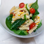 On a white table cloth, in a white bowl, mixed pasta salad, with spinach, fusili pasta, grape vine tomatoes cut in half, corn from a can