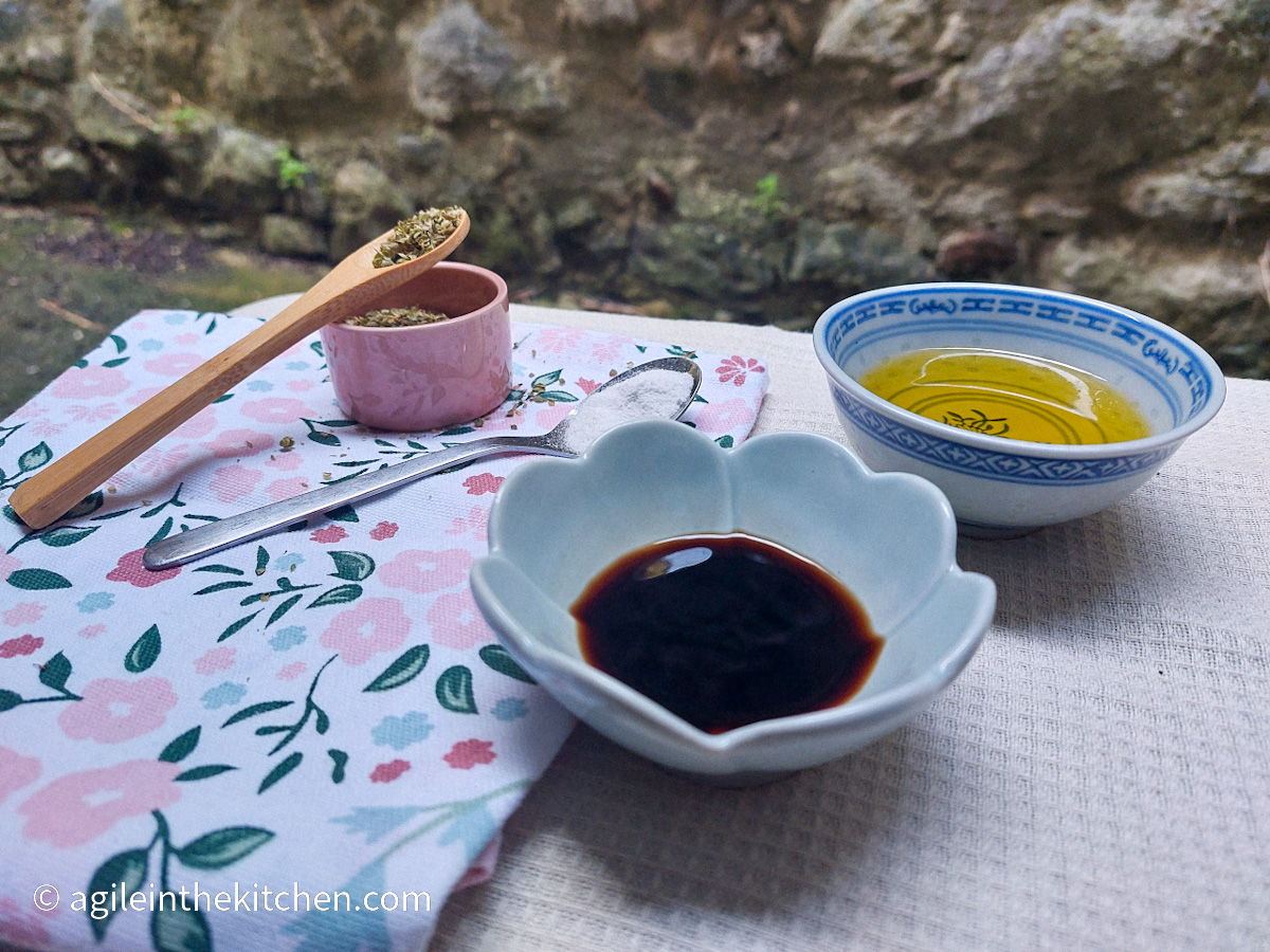 On a white table cloth, a folded cloth napkin with a flower and leaf pattern, in a small pink pot and a wooden spoon oregano, on a silver coloured spoon salt, a porcelain board with olive oil and a blue flower shaped bowl with balsamic vinegar.