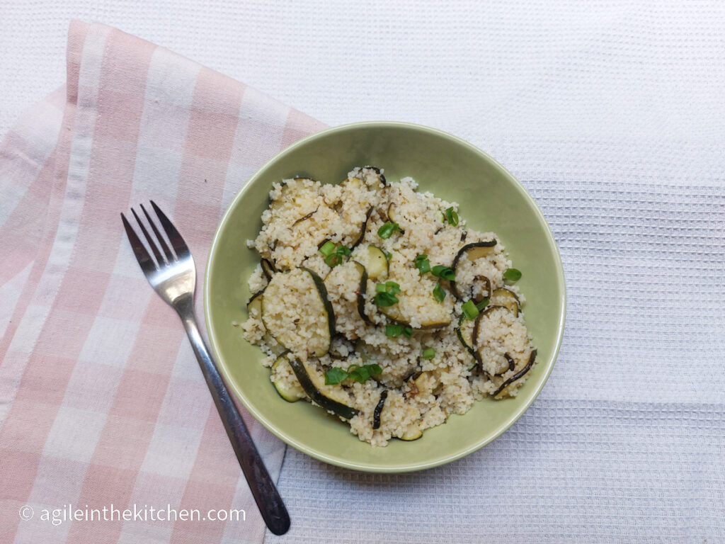 On a white background, a pink plaid folded cloth napkin, a fork, a green bowl with courgette and couscous salad topped with green onions