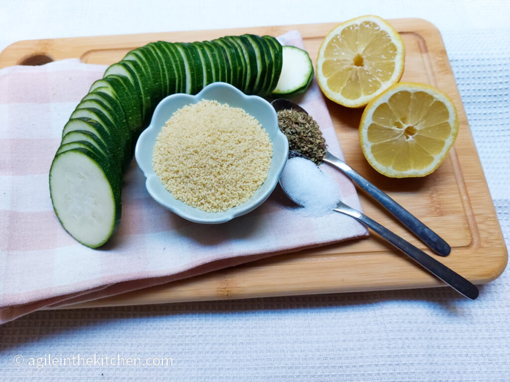 On a wooden cutting board, a pink plaid cloth napkin folded, then on top a courgette cut in thin slices, a flower shaped bowl with couscous, two teaspoons with salt and oregano, and a lemon sliced in half.