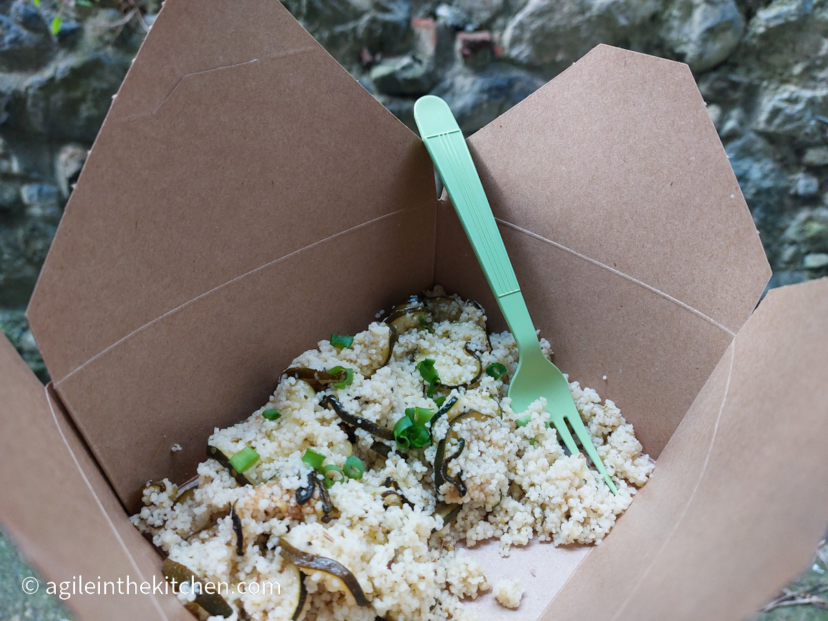 In a brown paper take out box, courgette and couscous salad and a green plastic fork.