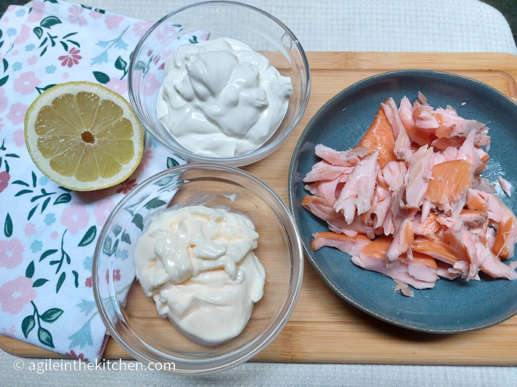 On a wooden cutting board with a flowery cloth napkin, the ingredients to make creamy salmon sauce. Clockwise, a lemon cut in half, a glass bowl of Greek yogurt, hot smoked salmon flaked on a blue plate, a glass bowl with mayonnaise