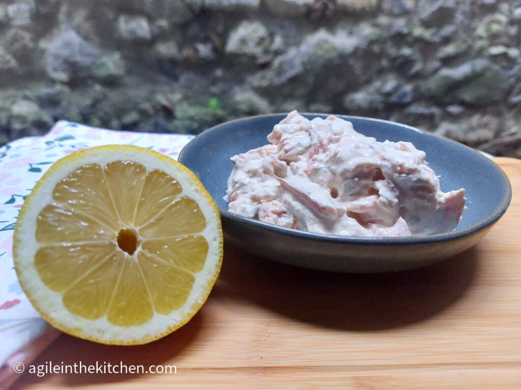 On a wooden cutting board, closest to the camera a lemon cut in half, in the middle a blue plate with creamy salmon mix, in the background a stone wall