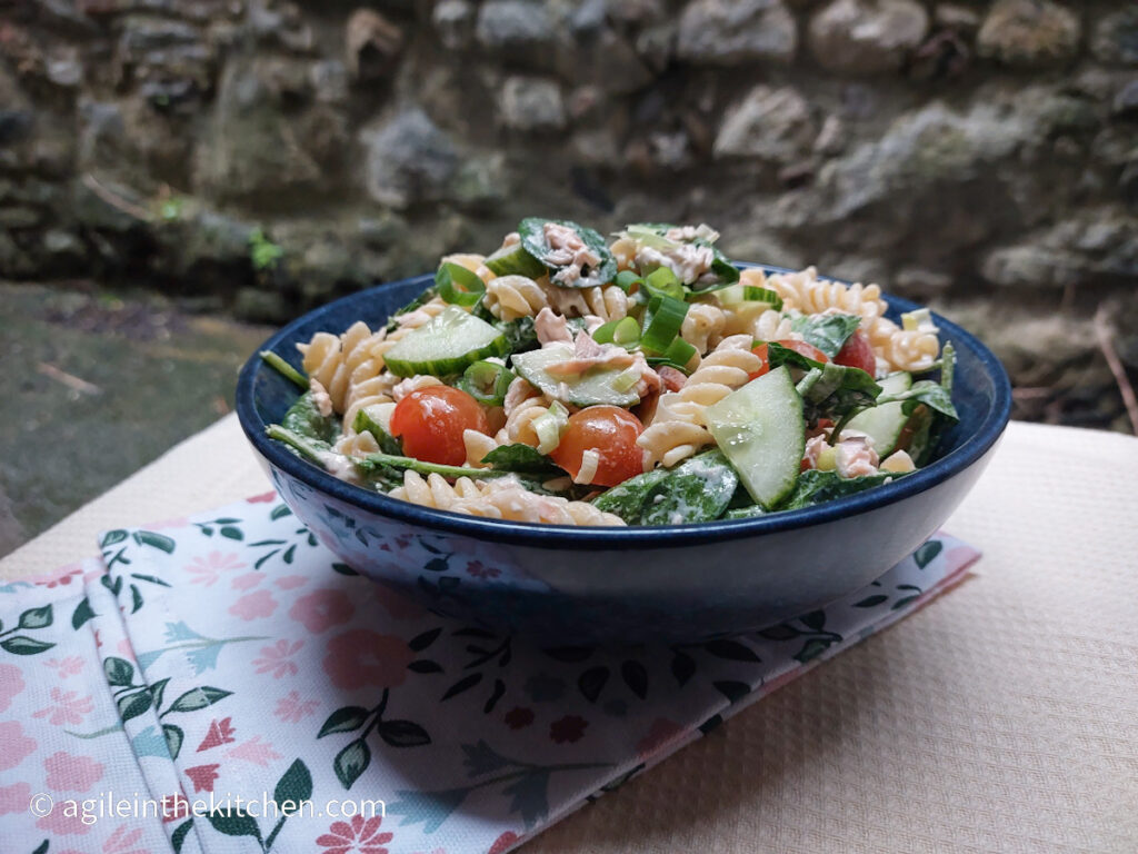 In a large blue bowl, creamy salmon pasta salad is mixed, visible are fusili pasta, spinach, cucumber, tomatoes cut in half and spring onions. On a background with a white, textured table cloth and a folded cloth napkin with pink and blue flowers.