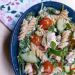 In a large blue bowl, creamy salmon pasta salad is mixed, visible are fusili pasta, spinach, cucumber, tomatoes cut in half and spring onions.