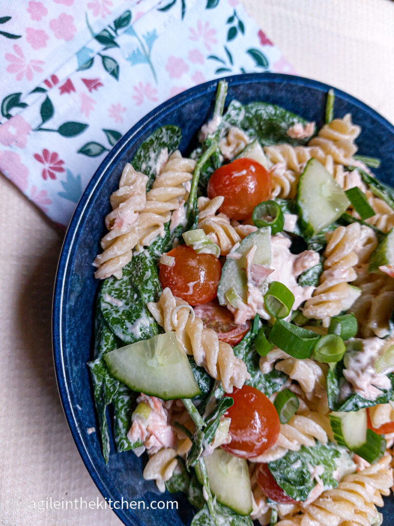 In a large blue bowl, creamy salmon pasta salad is mixed, visible are fusili pasta, spinach, cucumber, tomatoes cut in half and spring onions.