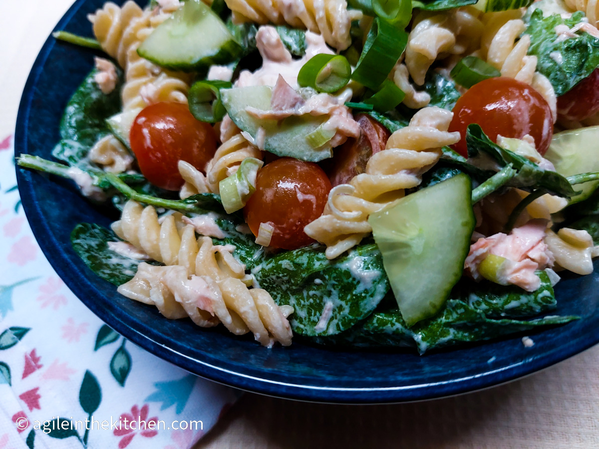 A close up of a large blue bowl with creamy salmon pasta salad. Visible are fusili pasta, spinach, cucumber, tomatoes cut in half and spring onions.