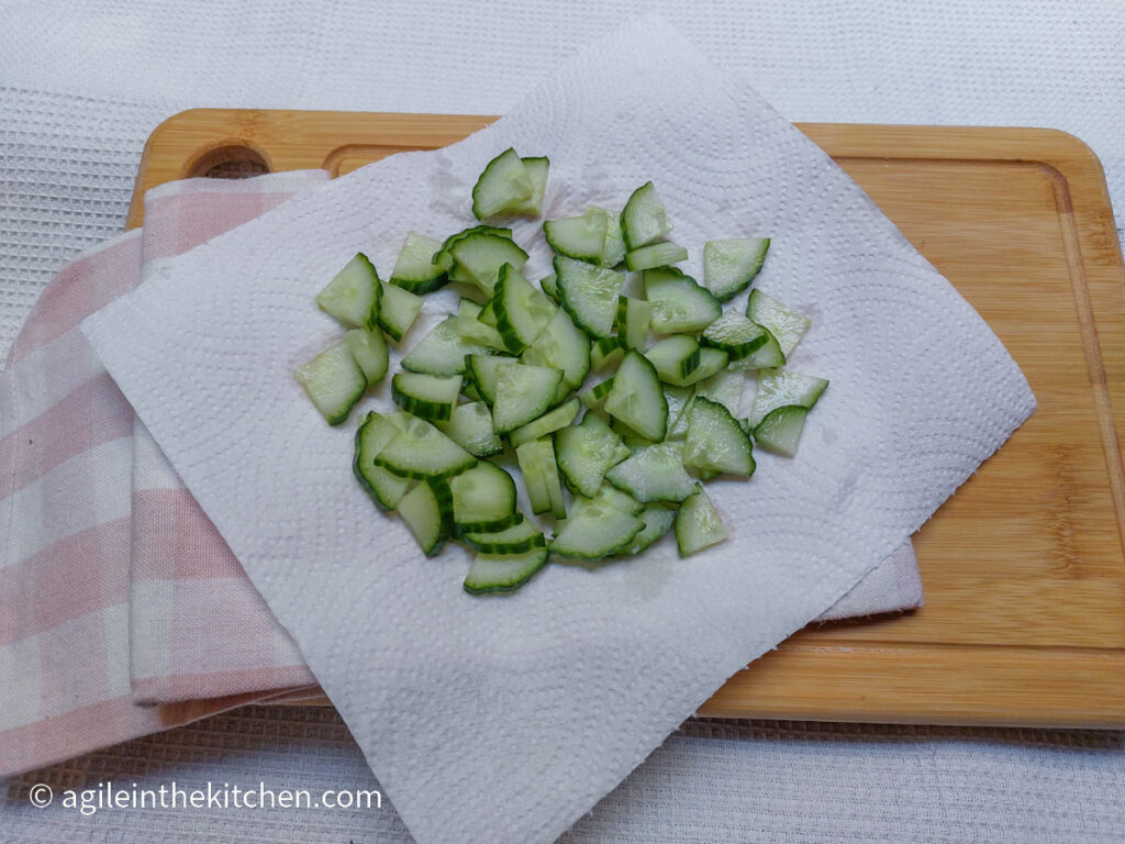 On a wooden chopping board with a pink plaid cloth napkin a piece of kitchen towel with cucumber pieces, sliced then quartered.