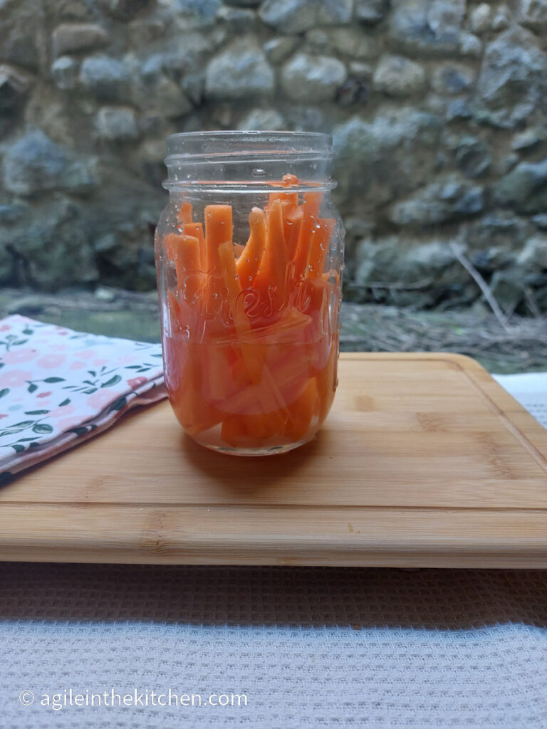 On a wooden cutting board, with a folded flowery cloth napkin, a mason jar with carrot sticks and pickle juice.